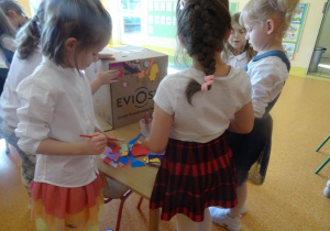 Trzy dziewczynki dekorują karton papierowy kolorowymi ścinkami papieru.